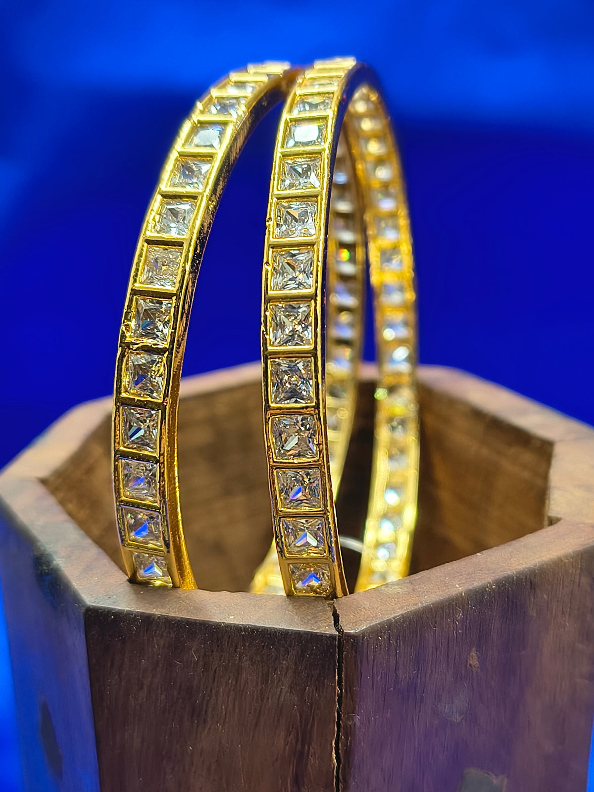 Two gold bangles adorned with square clear stones, displayed in a wooden holder against a vibrant blue background.